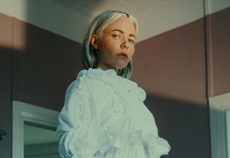 Kuvassa on Paula Vesala vaaleassa leningissä. Taustalla on ruskehtavaa seinää ja oven pieliä.