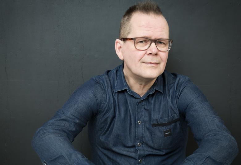 Kuvassa Kari Hotakainen puolivartalokuvassa istumassa tumman sinisessä paidassa  ja kuvan tausta on vielä tummempi harmaa.