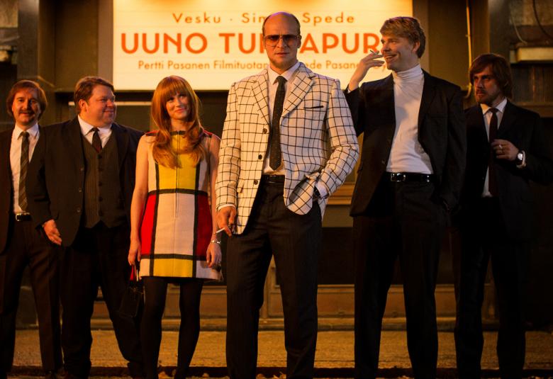 Kuvassa ovat elokuvan pääesiintyjät rivissä 1970-luvun asuissa ja hiusmuodissa.  Taustalla lukee Uuno Turhapuro.