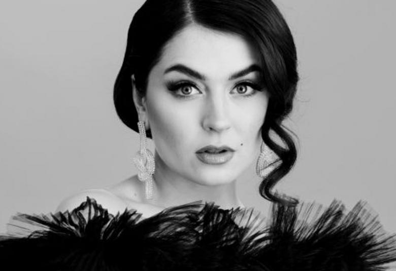  Kuvassa on BEHMin puolivartalokuva ja hänellä on avokaulainen röyhelöinen kaula-aukko mustassa asussaan.  Kuva on musta-valkoinen.