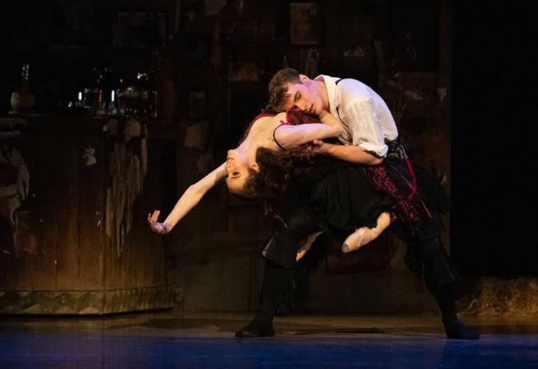 Kuvassa on tummalla näyttämöllä Rebecca King lähellä lattiaa taivutuksessa ja Hanako Matsune on kyykyssä ja pitää häntä sylissään.