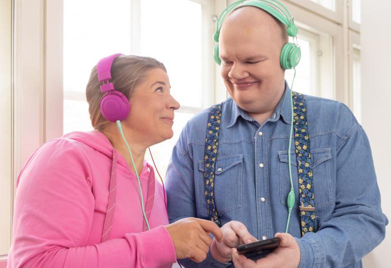 Kuvassa on keski-ikäinen nainen, joka katsoo oikealla puolellaan olevaa kookkaampaa miestä.  Naisella on vaalean punainen pusero ja violetit kuulokkeet päässään. Miehellä on vaalean sininen pusero ja vaalean vihreät kuulokkeet sekä kännykkä kädessään.