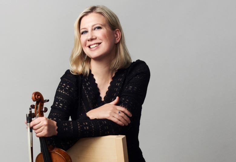 Kuvassa on Elina Vähälä istumassa tuolilla, jonka selkänoja on eteenpäin.  Hän Nojaa siihen ja pitää oikeassa kädessään viulua.  Kädet ovat ristikkäin.  Hänellä on musta pitsimäinen pusero päällään.