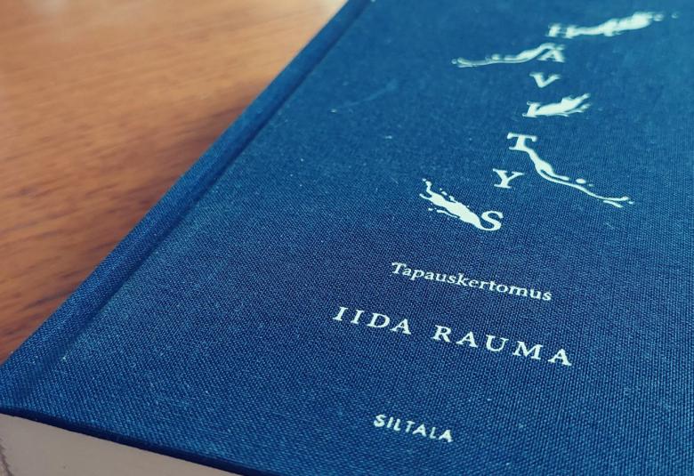 Kuvassa on sininen kirja vaalean ruskean pöydän kannella. Kirjan kannessa on sen nimi Hävitys -tapauskertomus valkoisella melko pienillä kirjaimilla, kirjoittaja Iida Rauma ja alhaalla kustantaja Siltala.