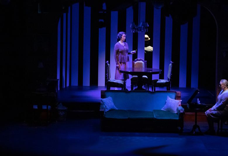 Kuvassa on kohtaus näyttämöltä ja siinä on sinertävällä näyttämössä, joka on olohuone. Siellä on ylempänä nainen seisomassa ja alempana toinen nainen istumassa. Kuvassa on runsaasti tummaa, jonka sisällä on kuva olohuoneesta.