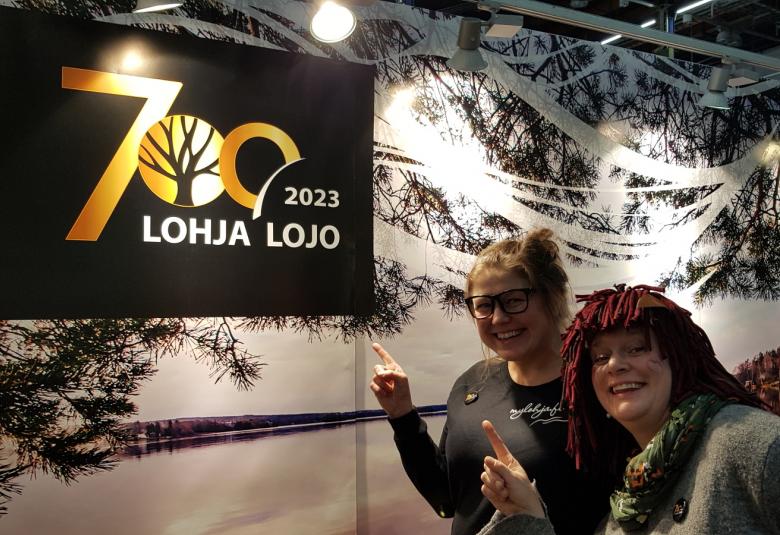 Kuvassa on vasemmalla yläkulmassa Lohja 700 vuotta juliste ja muu seinästä on Lohjanjärven maisemaa.  Oikealla ovat Jannika Joutsenniemi ja Tytyrin peikko osoittamassa julistetta.