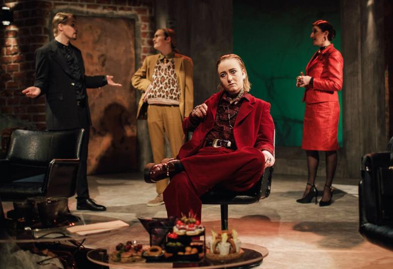 Kuvassa on ruskeasävyinen huone, jossa on reunoissa tummat pylväät ja tummia huonekaluja.  Takana on vasemmalla kaksi miestä keskustelemassa. Oikealla takana on punaisessa jakkupuvussa nainen ja etualalla on vaalea pöytä ja sen takana on nainen.