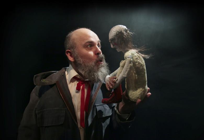 Kuvassa on Perttu Hallikainen, jolla on puoliksi paljas pää ja pitkät hiukset sivulla.  Kädessään hänellä on nukke.