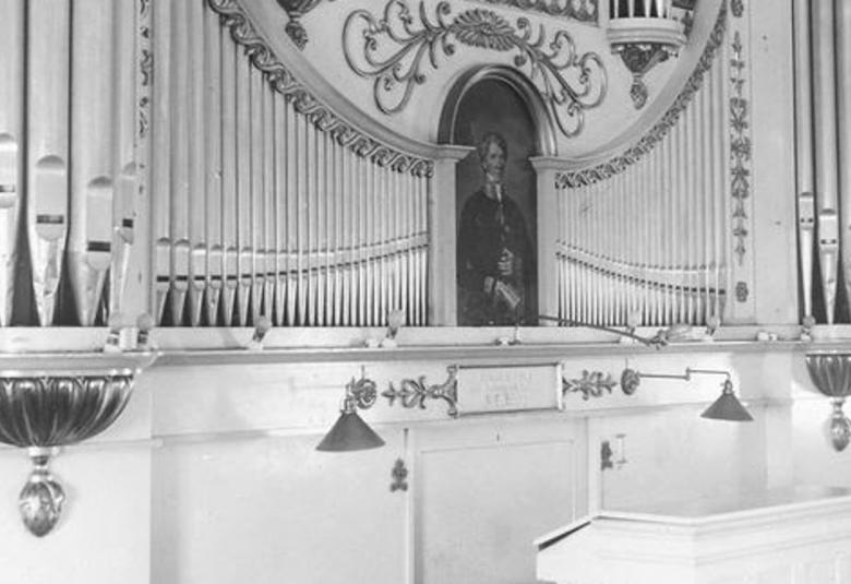 Kuvassa on osa Tammisaaren kirkon uruista ja niistä näkyy urkurin penkkiä ja urkupillejä sekä koristeellisia yksityiskohtia, joiden keskellä on taideteos.Kua on musta-valkoinen.