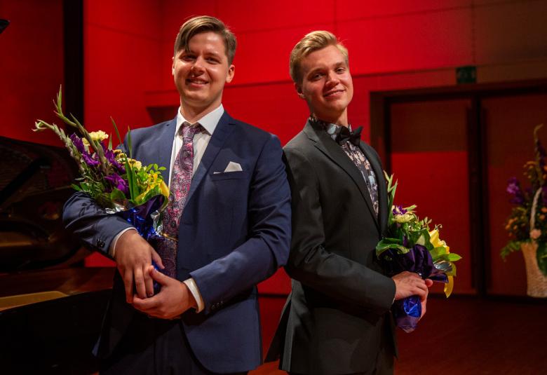 Kuvassa ovat Johan Krogius ja Jere Hölttä kukkakimput kädessään. He ovat puolivartalokuvassa ja tausta on punainen.