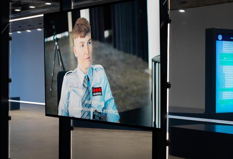 Kuvassa on pysty- ja vaakasuuntaisia mustia metallirunkoja ja niiden välissä on pleksit ja suuri vartijan kuva suuressa aulamaisessa tilassa. 