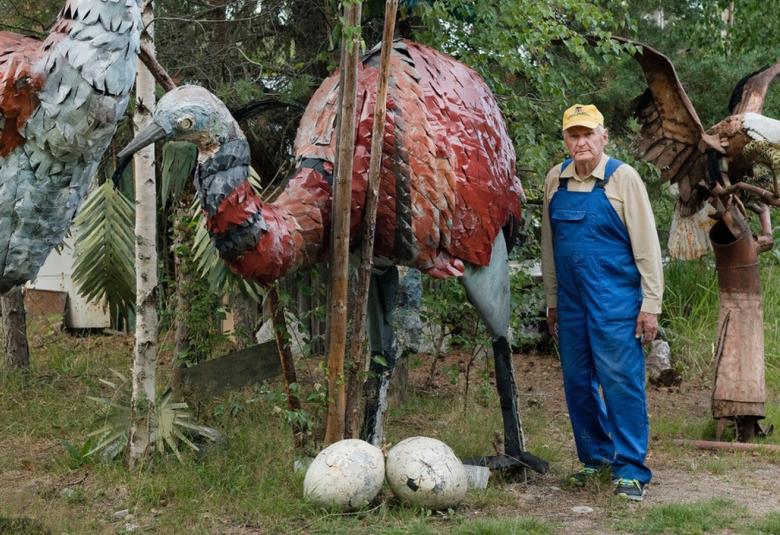Kuvassa on Alpo Koivumäki sinisessä haalarissa seisomassa suurten metallista tehtyjen isojen strutsien ja kannon päässä olevan kotkan keskellä. Maassa on kaksi isoa vaaleaa strutsinmunaa.