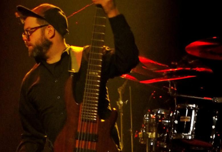 Kuvassa on soittaja sivukuvassa lippispäässä ja kitara kädessään.  Kuva on tumman sävyinen.