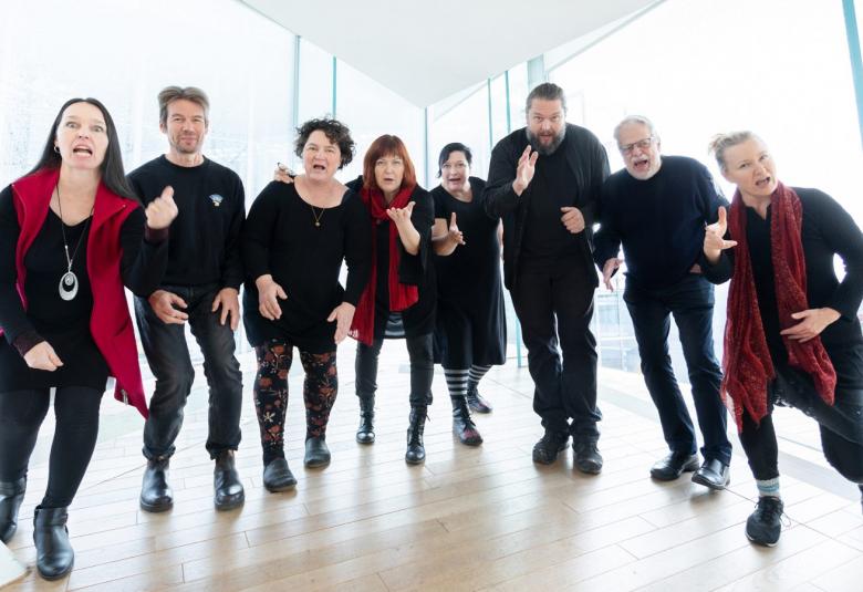 Kuvassa on Iki-Turson kahdeksan soittajaa, jotka ovat iloisilla ilmeillä hiukan eteenpäin kumartuneina valoisan konserttisalin aulatilassa.