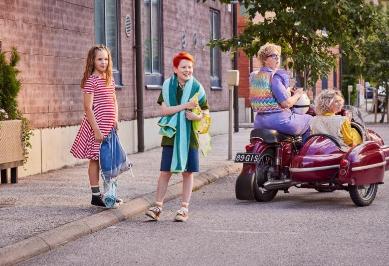 Kuvassa ovat Meri Lahikainen raitamekossa, Otso Törmälä vihreässä paidassa ja shortseissa sekä moottoripyörän sivuvaunussa poispäin menossa viininpunaisessa moottoripyörssä. 