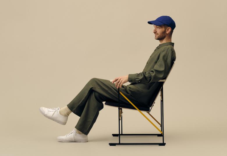 Kuvassa on Tomi Leppänen istumassa sivuttain rennosti ja hänellä on tumman sininen lippis päässään.  Tuoli on siro ja kevytrakenteinen.  Tausta on vaalean ruskea.