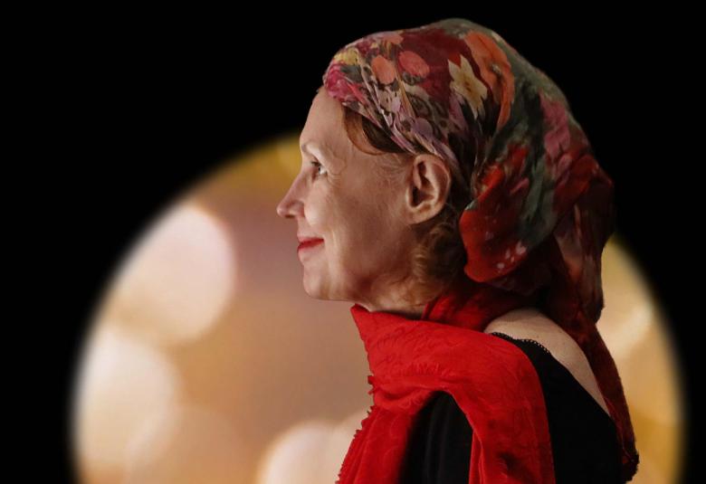 Kuvassa on kaija Saariaho sivuttain puolivartalokuvassa punainen huivi kaulassaan ja punakirjava huivi päässään löyhästi.  Taustalla on tummalla taustalla vaalean keltainen pyöreä kuvio.