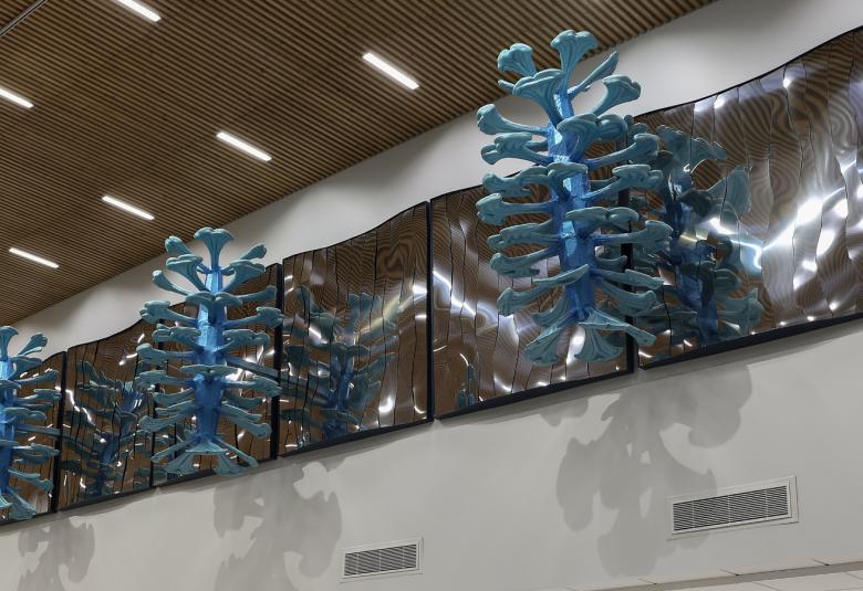 Kuvassa on lähellä ruskeaa sisäkattoa ja sen alla on valkealla seinällä tummalle taustalle kiinnitetty sinisiä puumaisia oksia ulospäin taustalevyistä.  oksat ovat taustalevyjen kokonaisia.