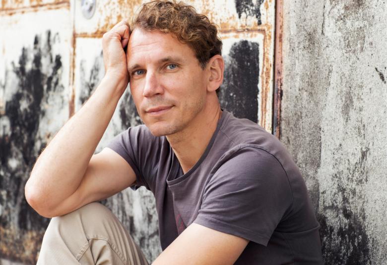 Kuvassa on Juha Itkonen istumassa lattialla ja nojaa oikealla kädellä polveensa ja poskeensa. Hänellä on harmaa t-paita ja vaaleat housut. Hänen taustallaan harmahtavaa kuvataidetta. 