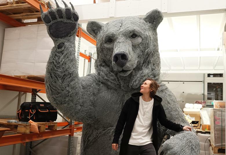 Kuvassa on Eliya Zweygberg ikäänkuin karhun kainalossa ja takana on harmaa karhuveistos.  Karhulla on käpälät pystyssä.