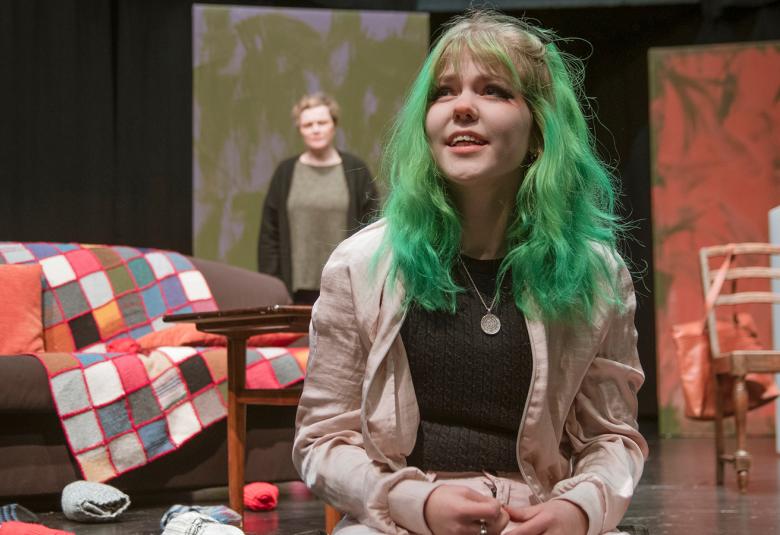 Kuvassa on nuori tyttö, jolla on pitkät vihreät hiukset ja vaaleat farkut. Hän istuu sohvalla ja taustalla näkyy huonetta sekä vanhempi nainen.