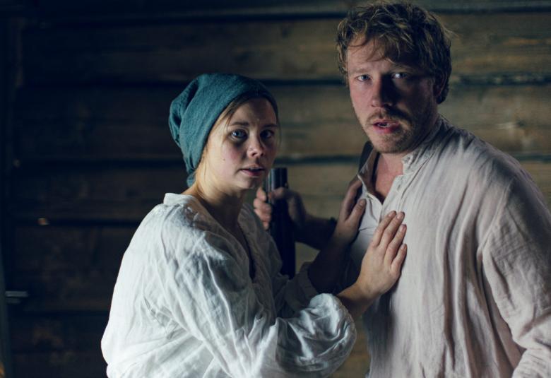 Kuvassa ovat Maija ja Janne ja heillä on vaaleat pellavapaidat päällään.  Kuva on puolivartalokuva ja takana näkyy tuvan hirsiä.