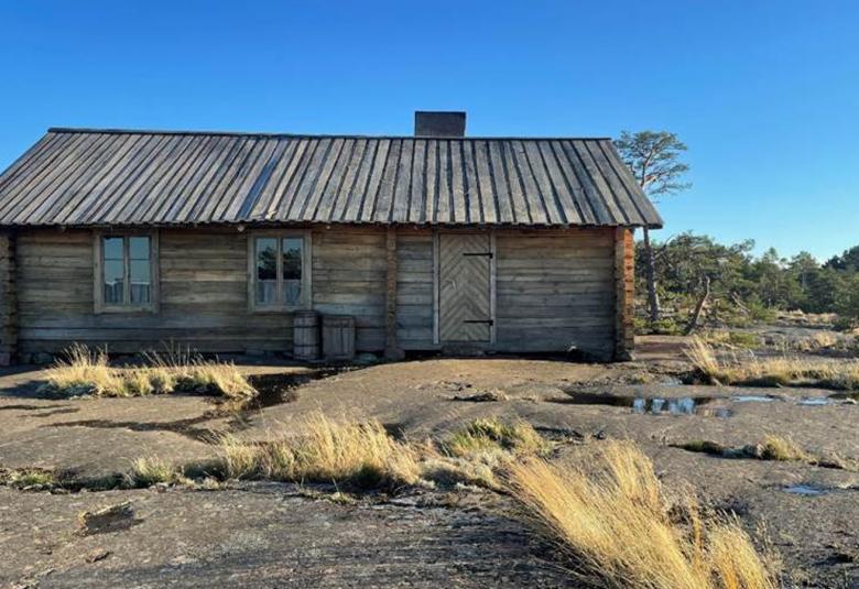 Kuvassa on kivisellä luodolla harmaa, vanha talo. Taustalla on sininen taivas.