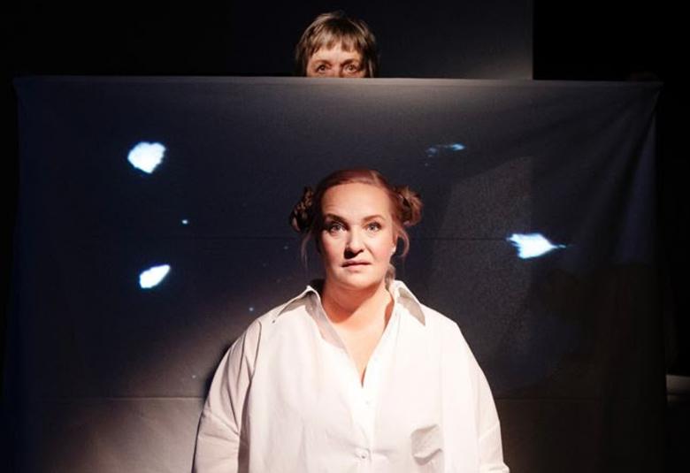 Kuvassa on tummalla taustalla eräänlaista kaksoiskuvaa, jossa on etualalla Nelly Juulia Kärkkäinen valkoisessa paitapuserossa. Takaa näkyy toinen kuva, josta näkyy puolet Gita Oksasen kasvoista.