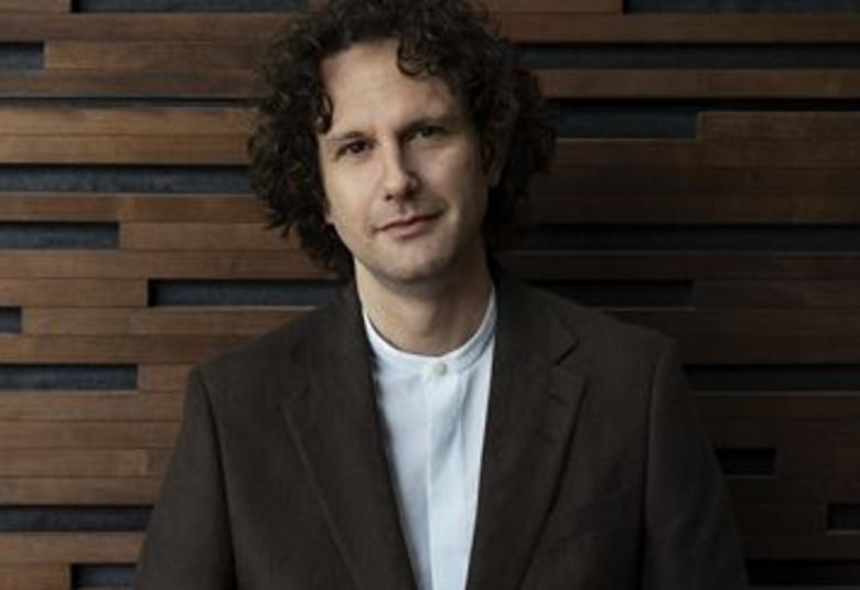 Kuvassa on Nicholas Collon, josta on puolivartalokuva. Hänellä on musta takki ja valkoinen paita päällään. Kuvan tausta on tumman ruskeaa puuseinää.