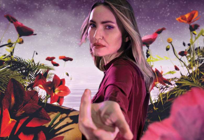 Kuvassa on nuori, pitkähiuksinen nainen, joka osoittaa vasemmalla kädellään kohti kameraa.  Hänestä on puolivartalokuva ja hänellä on lilan värinen pusero päällään.  Ympärillä on punaisia kukkia molemmin puolin.