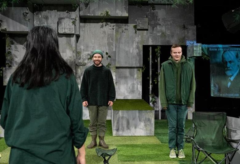 Kuvassa on takaperin Vilma Melasniemi ja taaempana seisovat parin metrin päässä toisistaan Juho Milonoff ja Miiko Toiviainen. Taustalla isojen tummien kivien seinää. Oikealla on valokuva vanhasta miehestä.