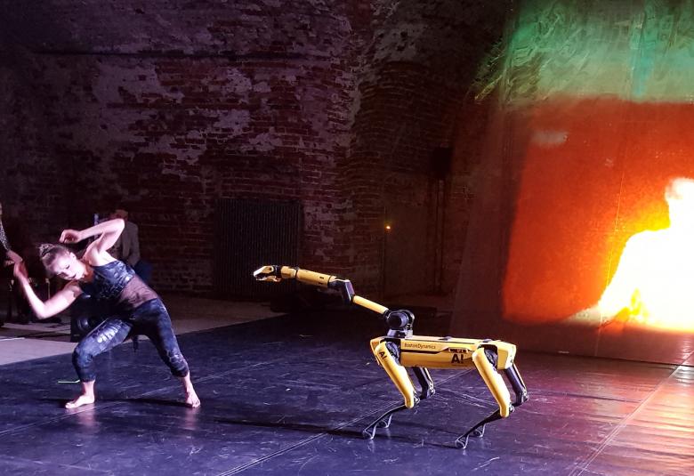 Kuvassa on tanssija väistämässä keltaista ison koiran kokoista keltaista robottia. Taustalla oikealla hologrammilakanassa tulen valoa.