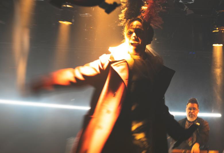 Kuvassa on Suvi-Sini Peltola ruskehtavassa takissa, johon osuu taustavaloa ja hän näkyy epätarkasti. Takana näkyy alhaalla kosketinsoittaja ja valopylväitä.