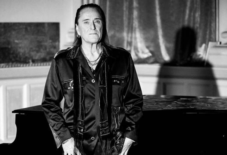 Kuvassa on Tommi Läntinen seisomassa huoneessa, jossa on takana flyygeli ja näkyy vaaleata, matalaa kaapistoa ja oikealla osittain teos seinällä.  Siinä on puun runkoja. Kuva on musta-valkoinen. 