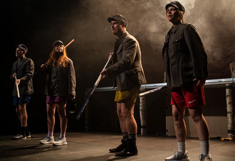 Kuvassa on näyttämöllä vinottain seisoo kaksi nuorta miestä shortseilla ja sotilasasissa ase kädessä.  Taaempana seisoo kaksi nuorta naista saanlaisissa asuissa.