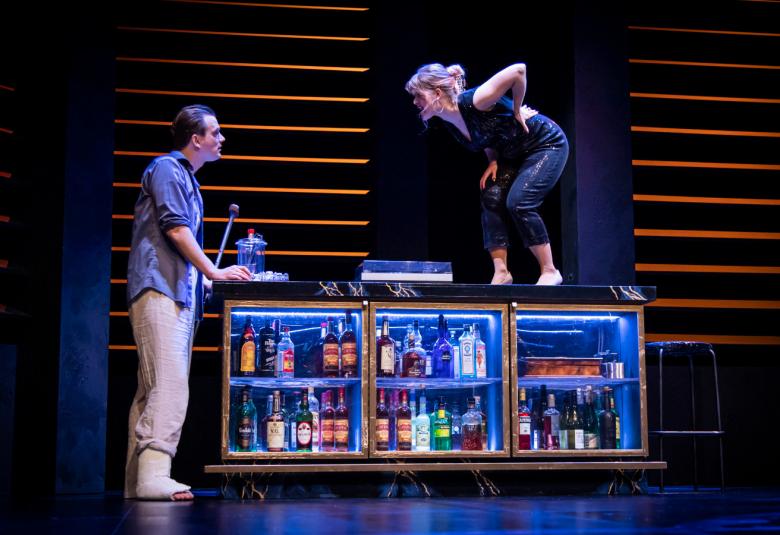 Kuvassa on vasemmalla mies oikean jalan nilkka kipsissä ja seisoo nojaten kaksihyllyiseen lasivitriinihyllyyn.  Nainen seisoo selkä taivutettuna hyllyn päällä. Tausta musta ja vaakalautainen.  Välissä on oranssi saumaus.