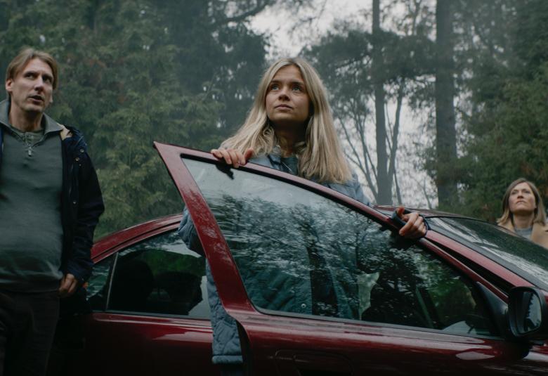 Kuvassa on vasemmalla Pekka Strang seisomassa ulkona tummassa ulkoilutakissa. Autosta on juuri nousemassa Saana Koivisto ja auton takana on Inka Gallén. Taustalla on metsää ja kuvan sävy on tumma.