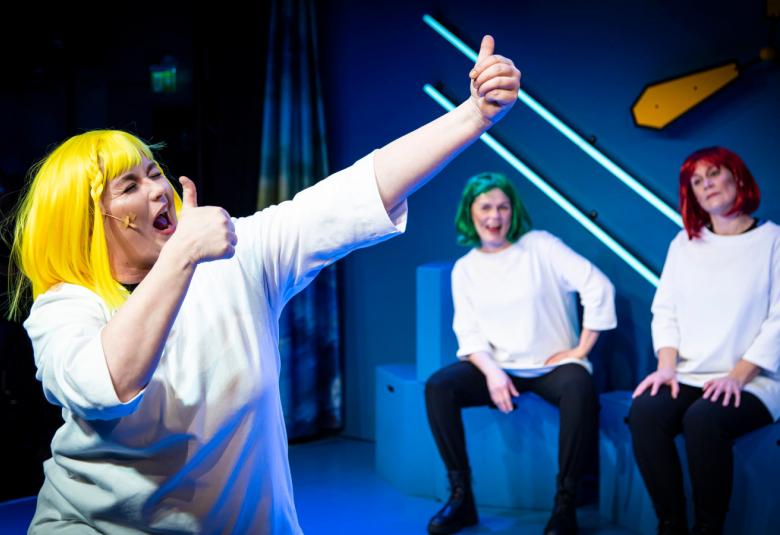 Kuvassa on naisnäyttelijä valkoisessa paidassa ja keltaisissa hiuksissa kädet vinosti ylöspäin ilmeikkäänä.  Taustalla kaksi naista istumassa.