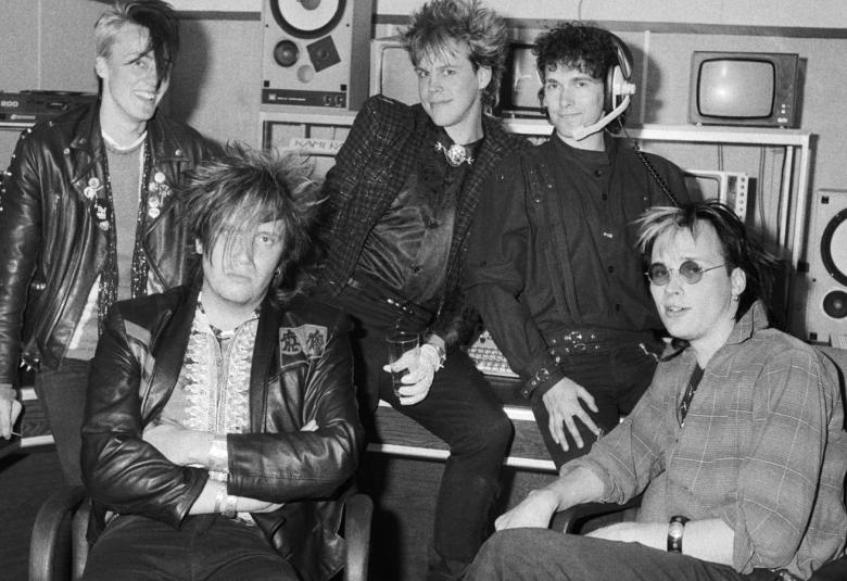 Kuvassa ovat Dingon viisi jäsentä bänditilassa ja kaksi jäsenistä istuu edessä tuoleilla ja muut ylempänä pöydällä.  Taustalla näkyy hyllyllä kaiuttimia. Kuva on musta-valkoinen.