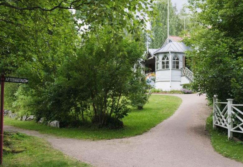 Kuvassa on Domanderin talo, joka näkyy vehreiden puiden lomassa ja sinne johtaa kapea hiekkatie ja toinen etualalla oleva hiekkatie. Talo on puusta ja harmaaksi maalattu.  Siitä näkyy kulmikas erkkeri. 