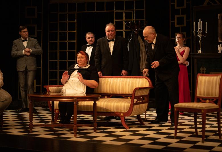 Kuvassa on seitsemän henkilöä katseet sohvalla istuvaan mustassa mekossa ja valkoisessa essussa olevaan ja sohvalla istuvaan palvelijanaiseen.  Kalusteet ovat vanhanaikaisia.