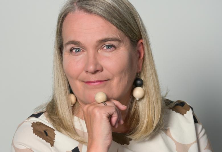 Kuvassa on Leena Valmu, josta on puolivartalokuva ja hän nojaa toiseen poskeenssa.  Hänellä on vaaleat puolipitkät hiukset ja päällään vaalean ruskea Unikko-kuosinen pusero päällään.
