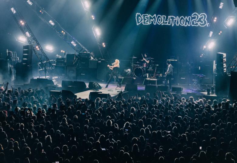 Kuvassa on lavalla esiintymässä Michael Monroe Demolition 23 -yhtyeen kanssa ja nimi on isolla taustalla. Etualalla on yleisöä.  Kuvan sävy on sininen.