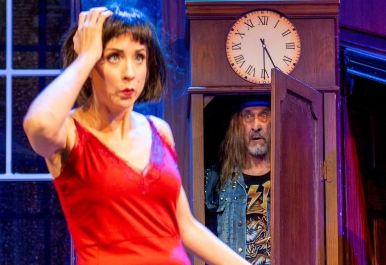 Kuvassa on nuori nainen punainen hihaton mekko päällään. Takana on kaappi, jossa on sisällä parrakas mies.