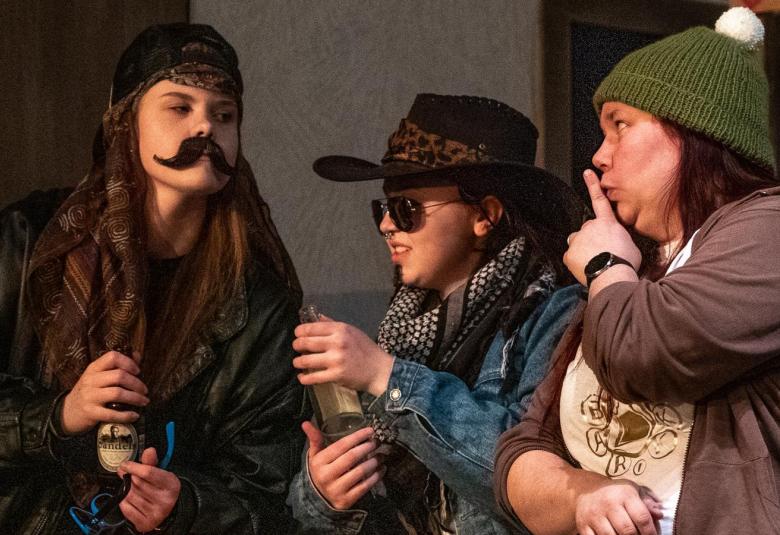 Kuvassa on kolme nuorta baaripöydän ääressä. Vasemmalla on nuorta miestä esittävä nuori nainen, jolla on musta lippis, musta nahkatakki ja kädessä olutpullo.  Keskellä on nuorimies, jolla on stetson päässään ja sininen farkkutakki. Oikealla reunassa istuu nuori nainen, jolla vihreä pipo ja ruskea villatakki.