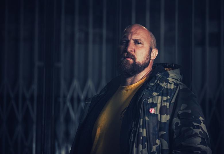 Kuvassa on Paleface oikealla puolella puolivartalokuvassa ja hänellä on tummakuvioinen pikkutakki. Toinen puoli kuvasta on tumma.