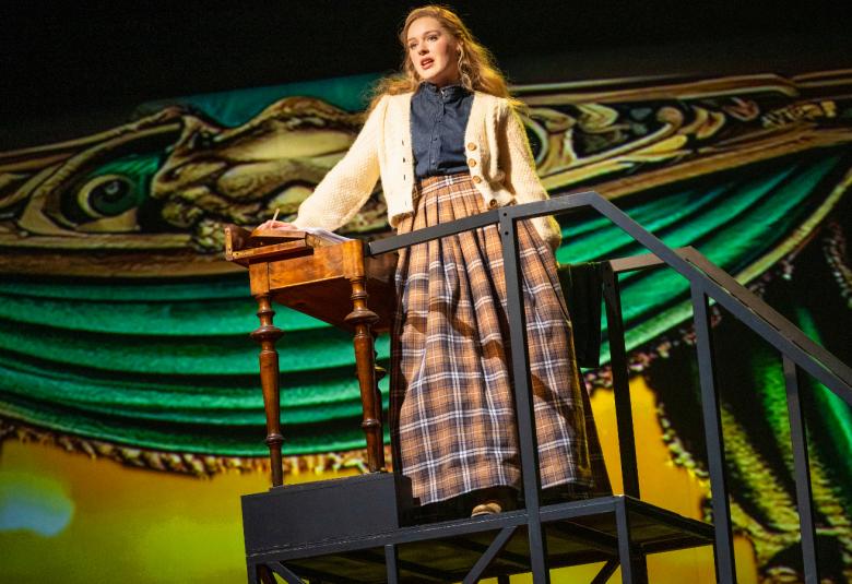 Kuvassa on Isla Mustanoja yläviistoon kuvattuna ja hän seisoo ylhäällä olevalla pienellä mustalla parvekkeella. Hänellä on pitkä, ruudullinen puolihame ja vaalea villatakki.  Taustalla on vihreitä soikeita valokuvioita.