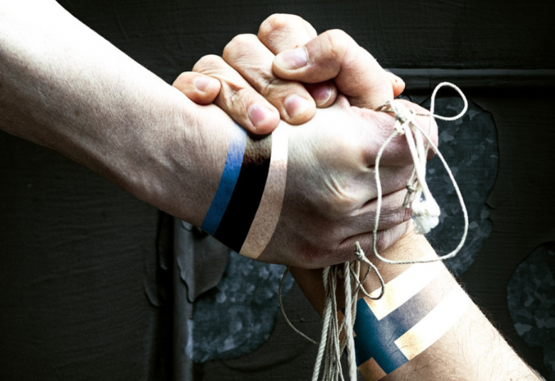 Kuvassa on kaksi eri henkilön kättä, joiden nyrkit ovat puristuneet yhteen.  Toisell on kädessään Suomen lippu ja toisessa Viron lippu.