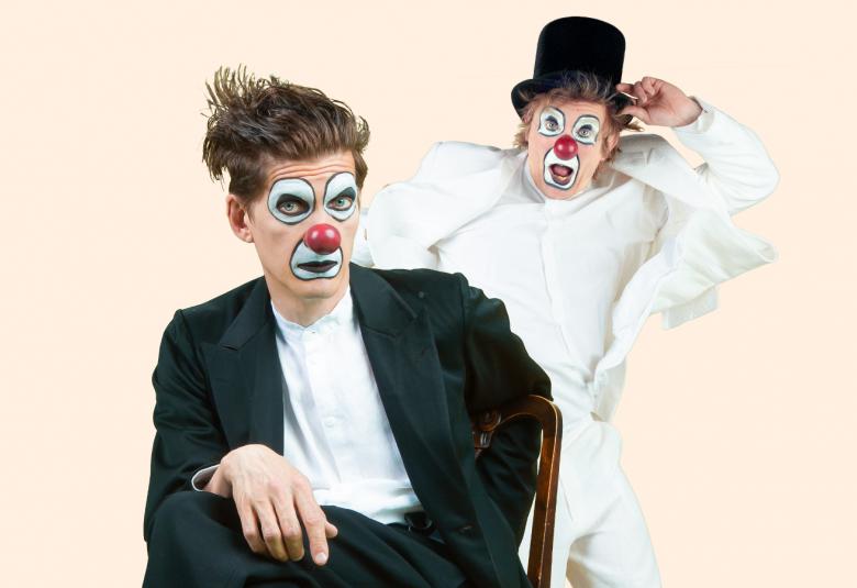 Kuvassa on etualalla Timo Ruuskanen sirkusmaisessa maskissa istumassa mustassa puvussa ja takana Tuukka Vasama vastaavalla maskilla ja vaaleassa asussa.