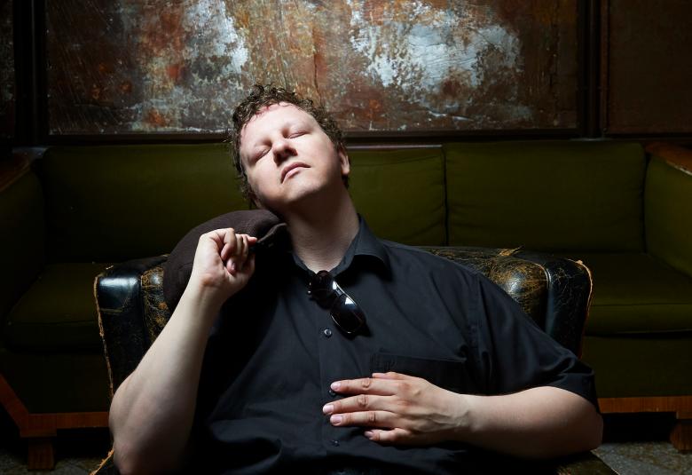 Kuvassa on Kalle Salonen, joka istuu hiukan takaviistosti ja ojentaa vasenta kättään eteenpäin.  Kuvan sävy on tumma.